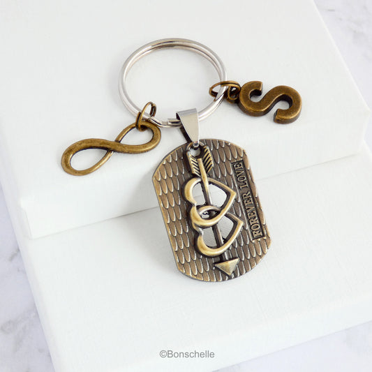An antique bronze toned keyring with a focal charm featuring two hearts and an arrow and the words 'forever Love', pluse an optional initial charm and an infinity sign charm.