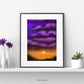 Original Art Poster showing an orange and purple sky with a setting sun mounted on a white background with a black frame, sitting on a white shelf with flowers in vases and a pot each side