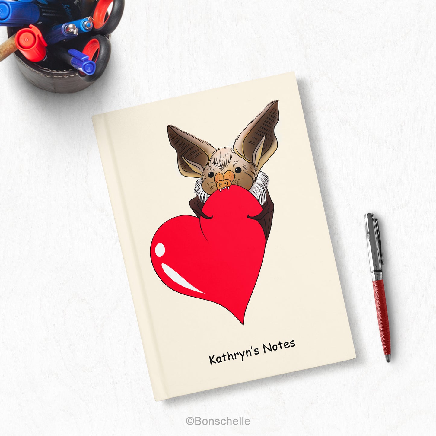 Cute Personalised Hard Cover Notebook, with a Chibi Bat holding a red love heart and personalisation on the front shown on a desk