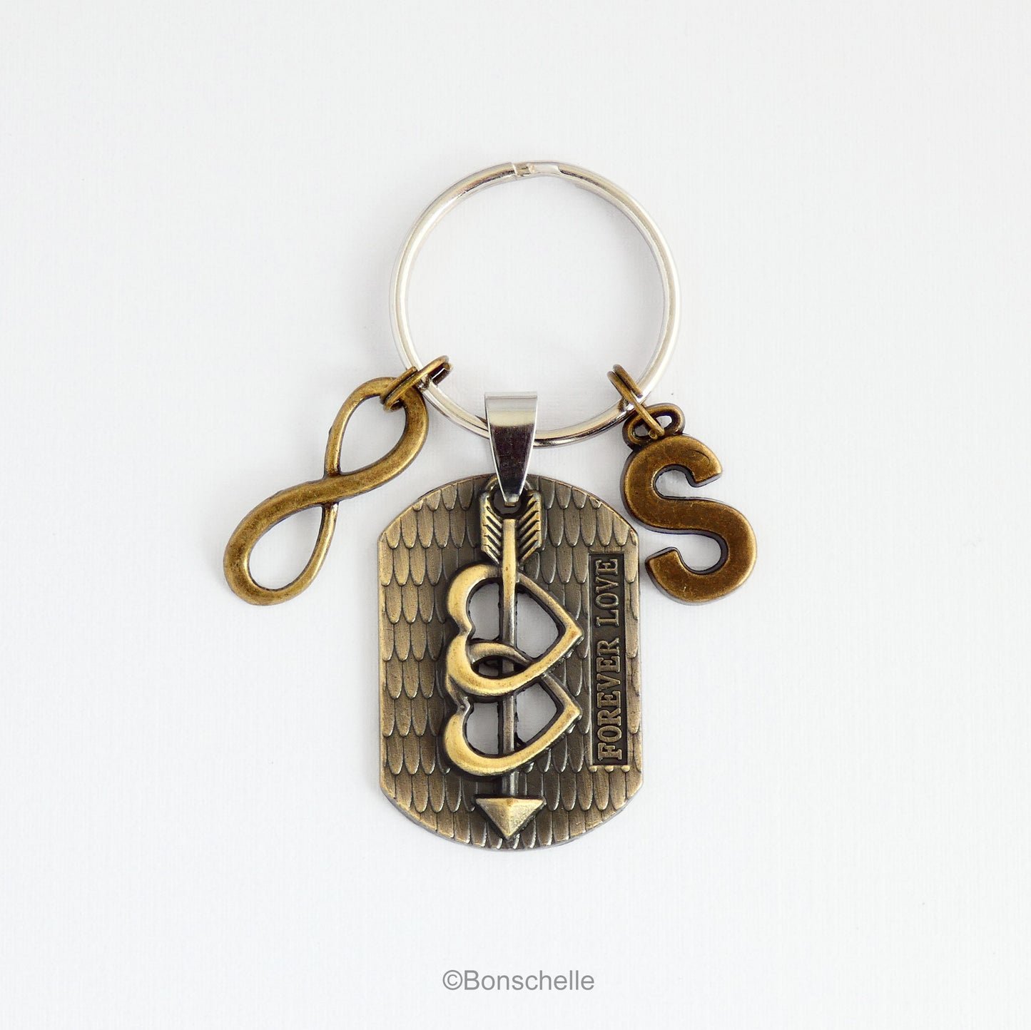 An antique bronze toned keyring with a focal charm featuring two hearts and an arrow and the words 'forever Love', pluse an optional initial charm and an infinity sign charm.