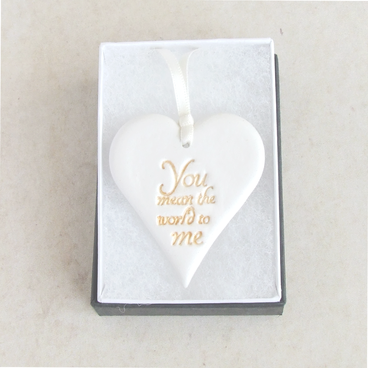 Heart shaped hanging ornament made from white clay with gold words 'You mean the world to me' , laying in a gift box.