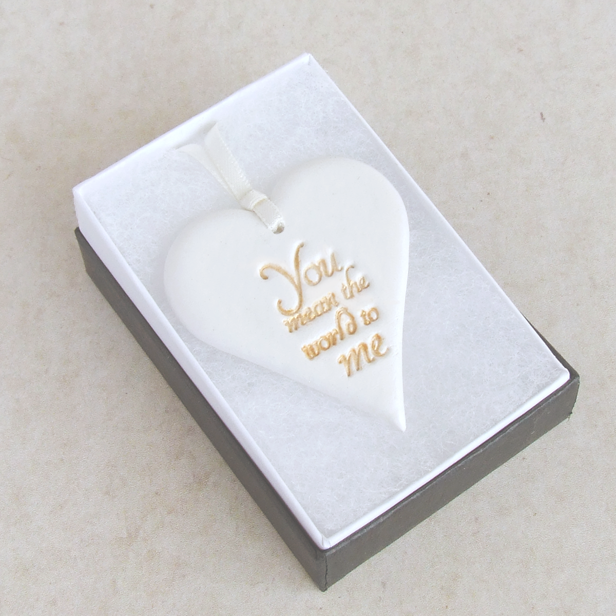 Heart shaped hanging ornament made from white clay with gold worlds 'You mean the world to me' , laying in a gift box.