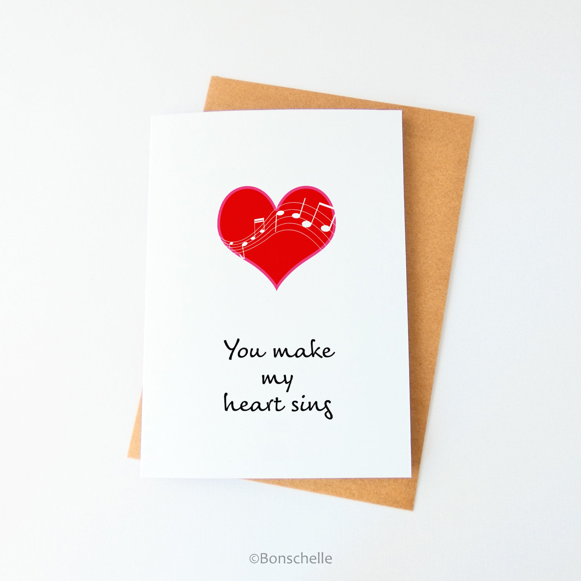 Front view of a handmade white greeting card with a red heart with white musical notes overlaid, and the words You Make My Heart Sing beneath, with a brown envelope.